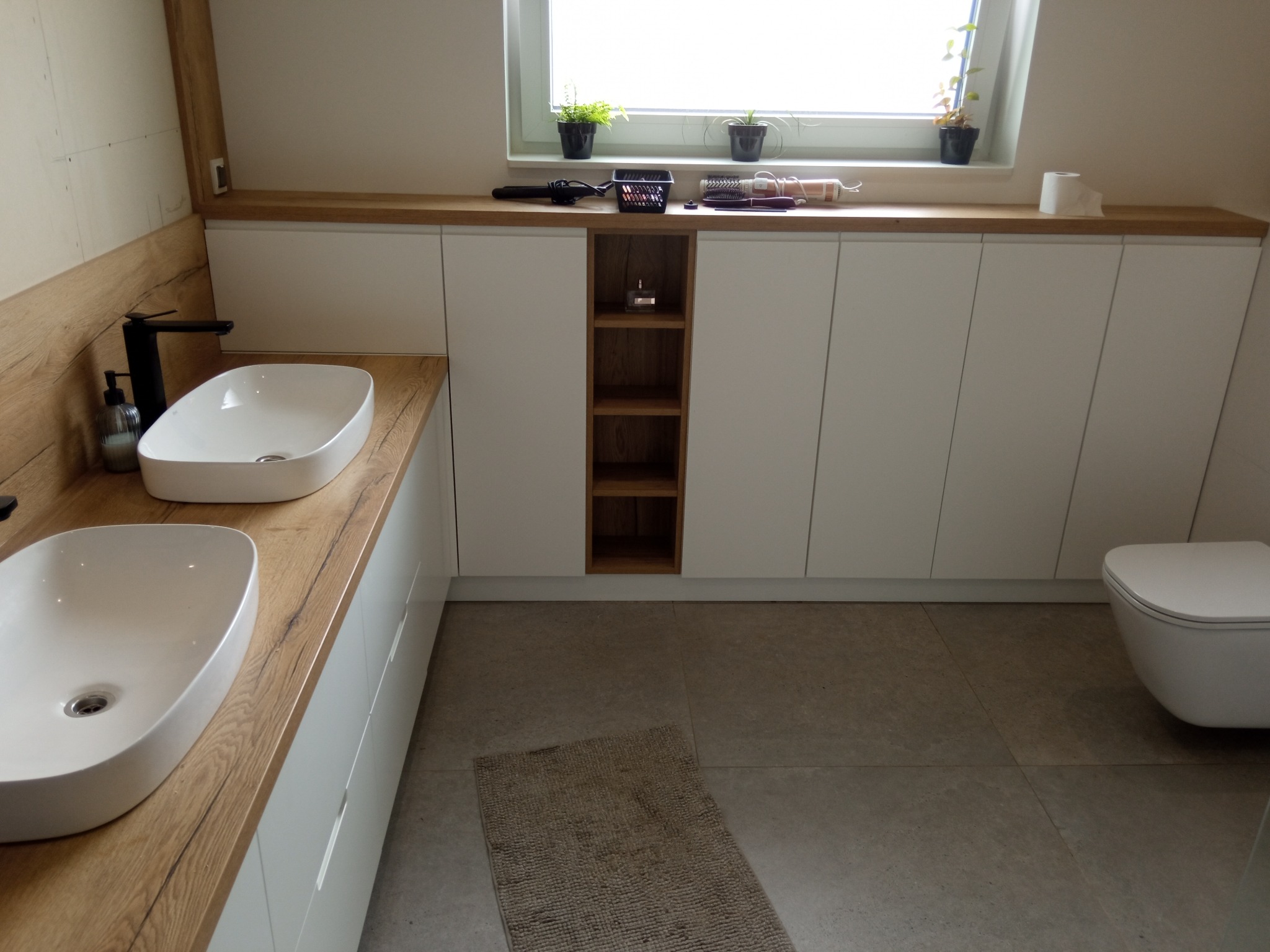 Nowoczesna łazienka z dwoma umywalkami nablatowymi na drewnianym blacie, białymi szafkami podblatowymi z otwartą półką, oświetlonym oknem i beżową podłogą.