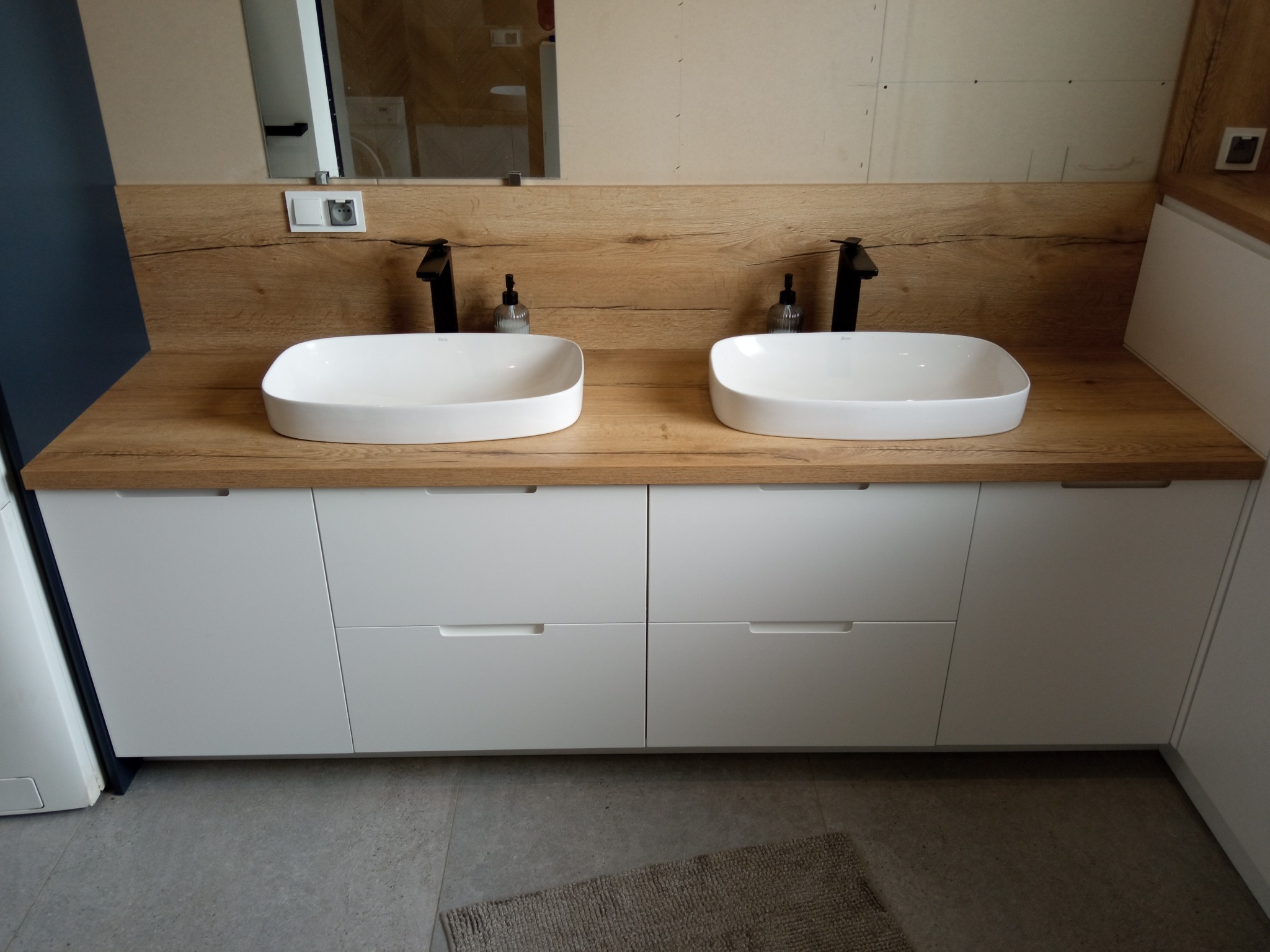 Nowoczesna łazienka z dwiema białymi umywalkami nablatowymi, czarne baterie, drewniany blat i białe szafki pod umywalką.