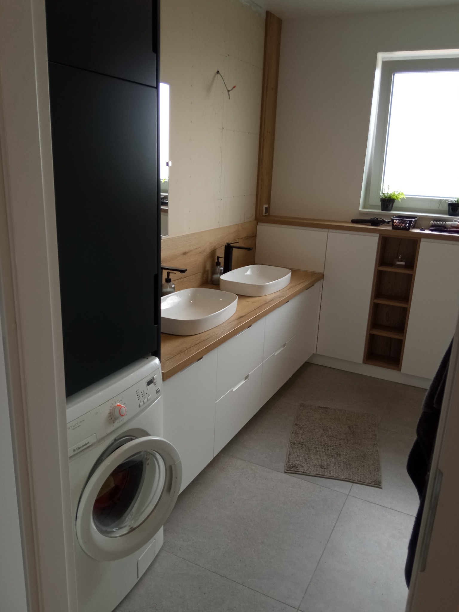 Nowoczesna łazienka z dwoma umywalkami nablatowymi, drewnianym blatem, białymi szafkami z szufladami i czarnym wysokim regałem nad pralką.