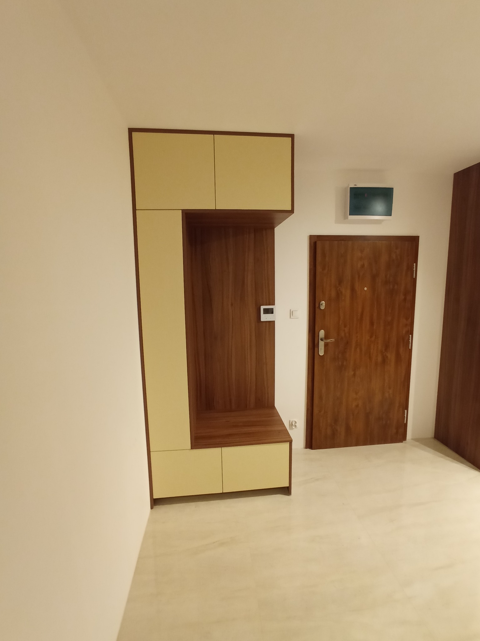 Nowoczesny przedpokój z zabudową meblową w odcieniach beżu i drewna, obejmującą szafki górne i dolne oraz otwartą wnękę z półką, obok drewniane drzwi wejściowe.