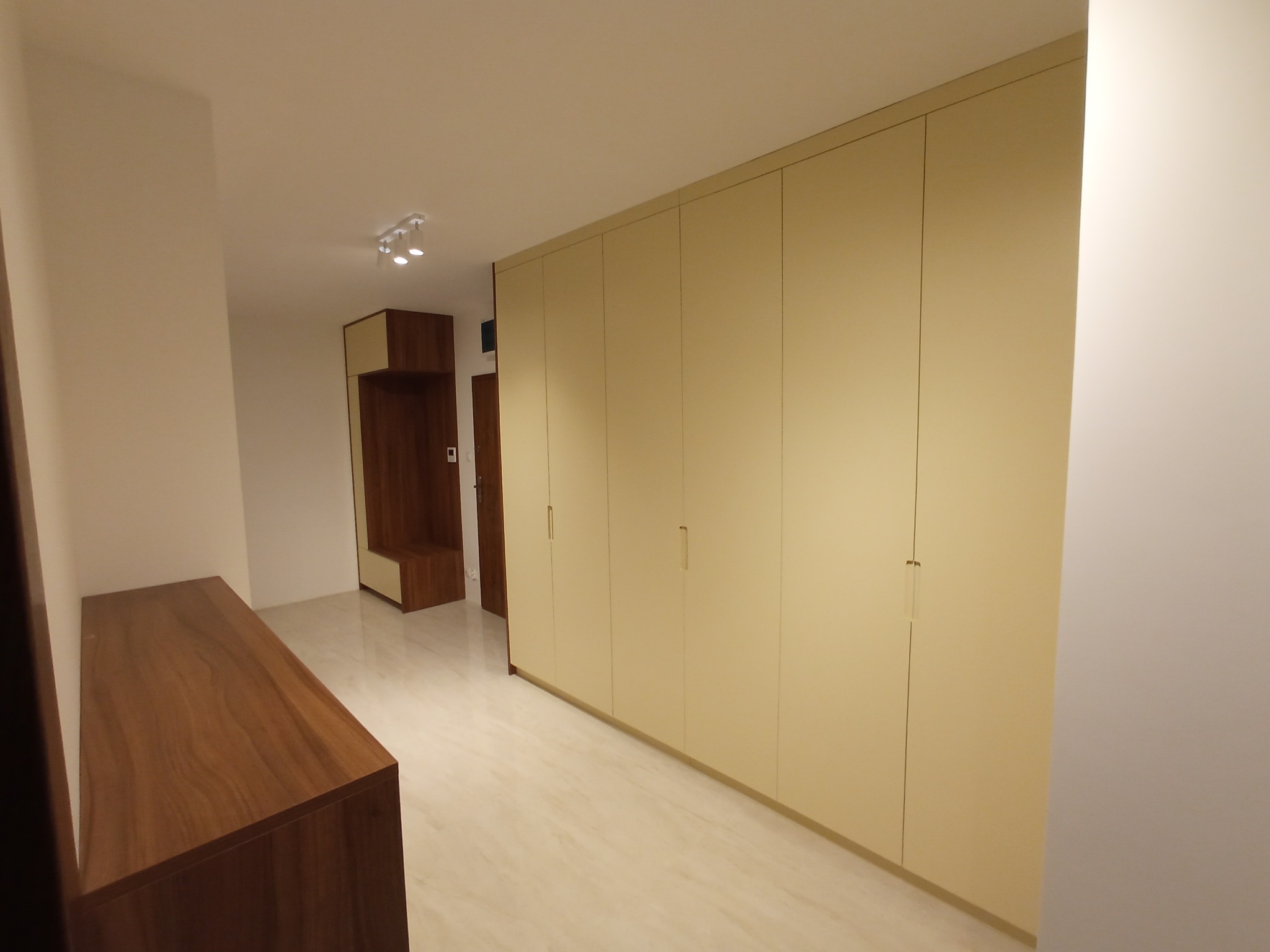 Długa, beżowa szafa wnękowa z minimalistycznymi uchwytami, zabudowana od podłogi do sufitu, widoczna w korytarzu z jasną podłogą i drewnianą komodą.