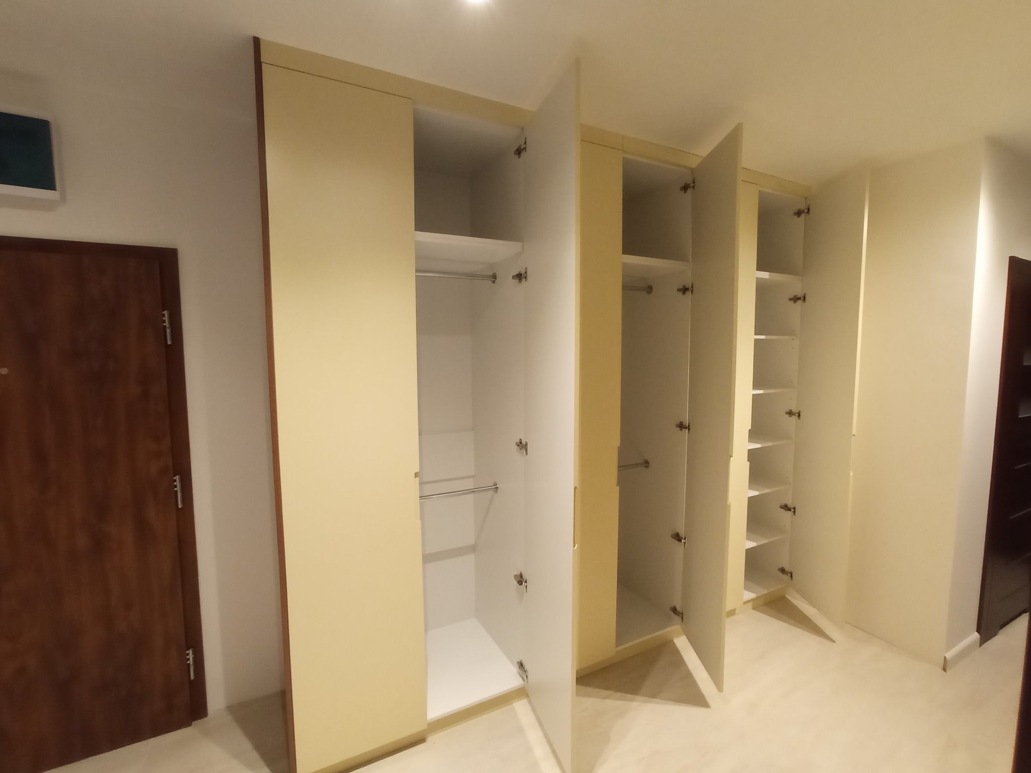 Beżowa szafa wnękowa z otwartymi białymi drzwiami, widoczne półki i drążki na ubrania, zamontowana w jasnym pomieszczeniu z drewnianą podłogą i drzwiami.