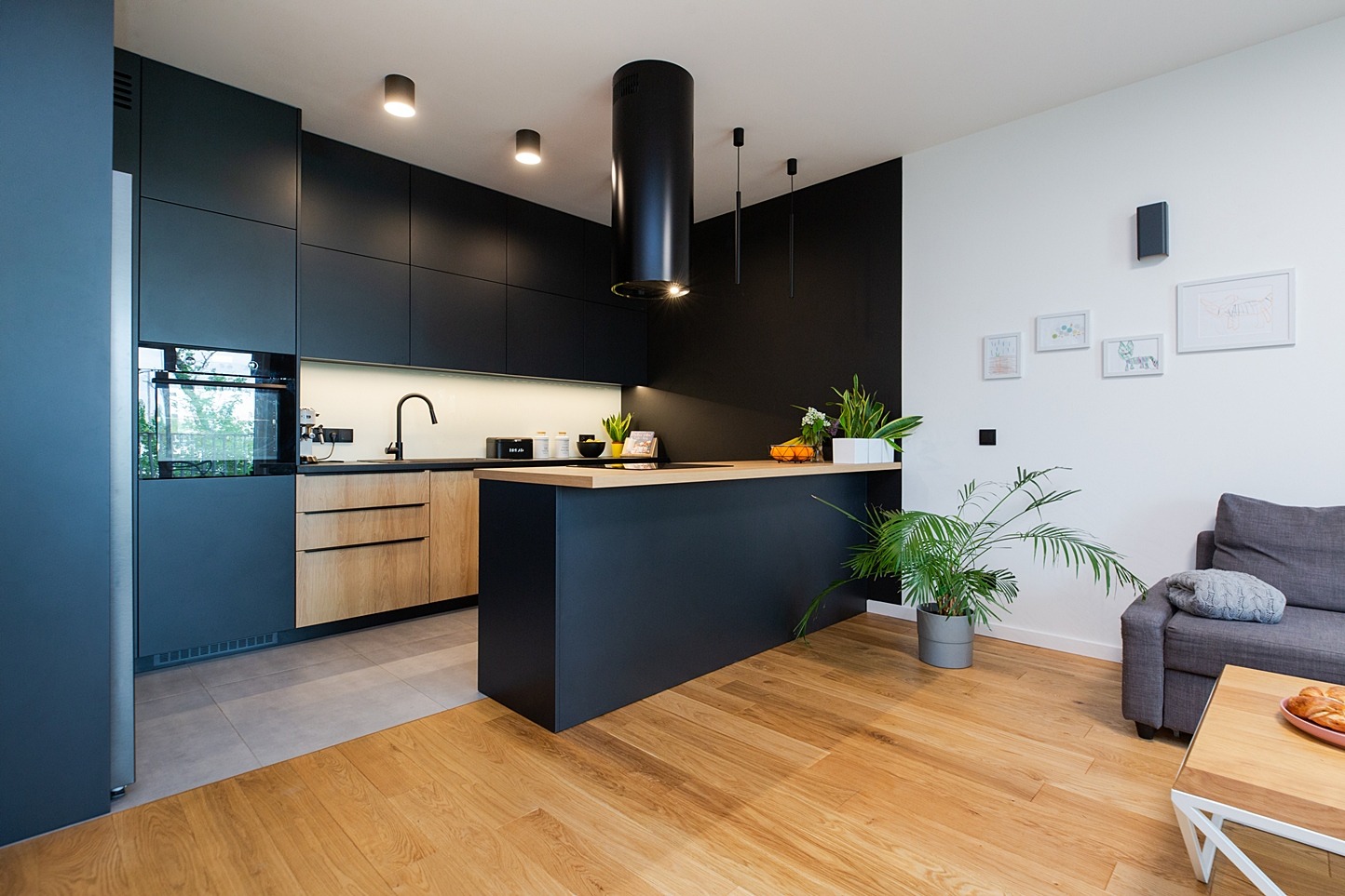 Nowoczesna kuchnia z czarnymi szafkami, drewnianymi frontami dolnymi, wyspą z blatem, okrągłym okapem i drewnianą podłogą, widok na salon z szarą kanapą.