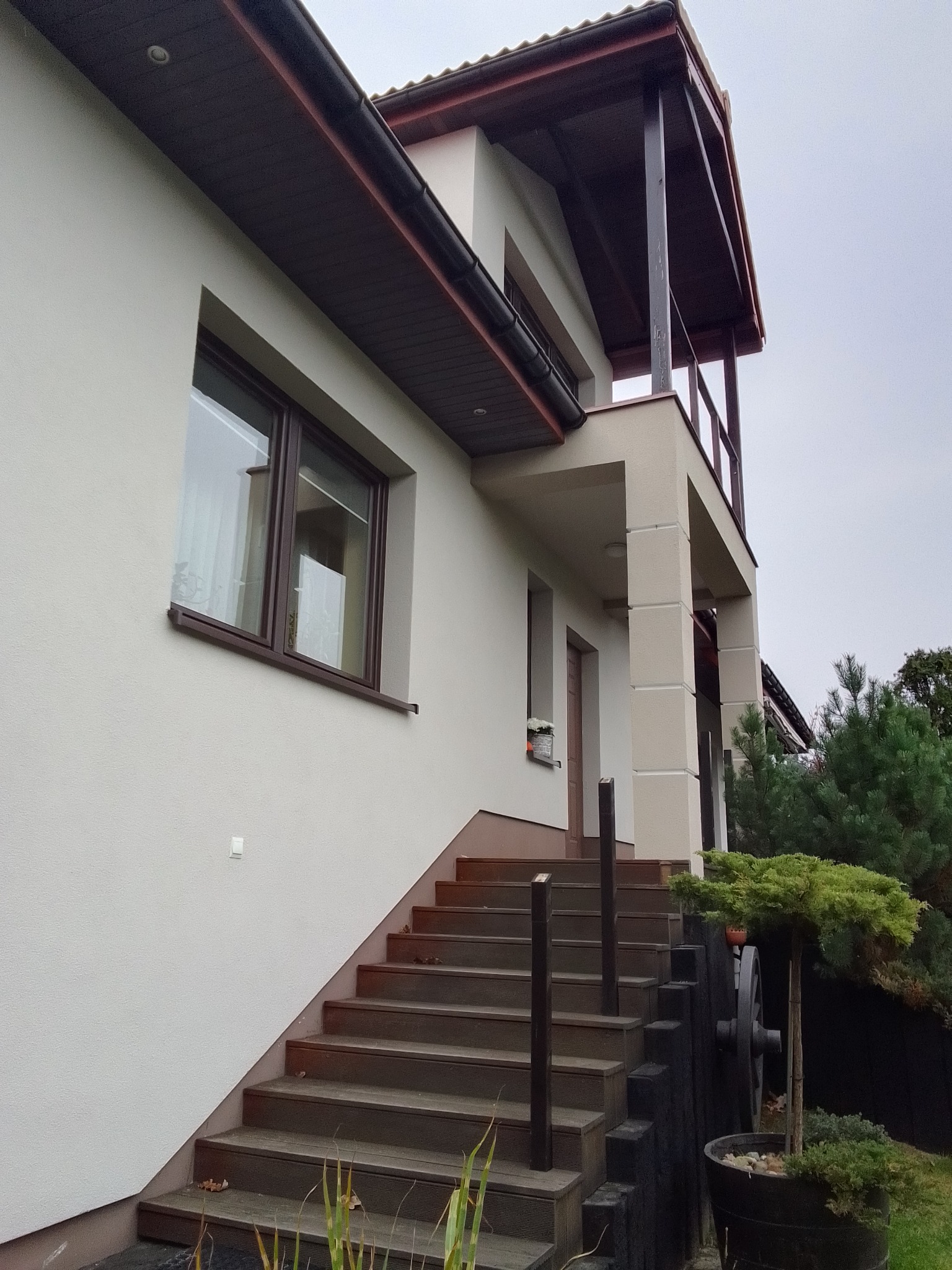 Elewacja domu jednorodzinnego w jasnych barwach z ciemnobrązowymi elementami wykończeniowymi, schody wejściowe z drewnopodobnych desek, dekoracyjny wózek z kołem i małym drzewkiem w donicy.