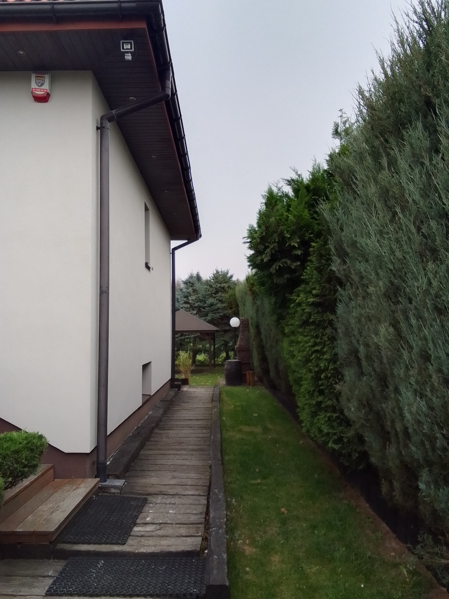 Boczny widok otynkowanego domu z brązowymi rynnami, drewnianym chodnikiem i zielonym żywopłotem, w tle altana.