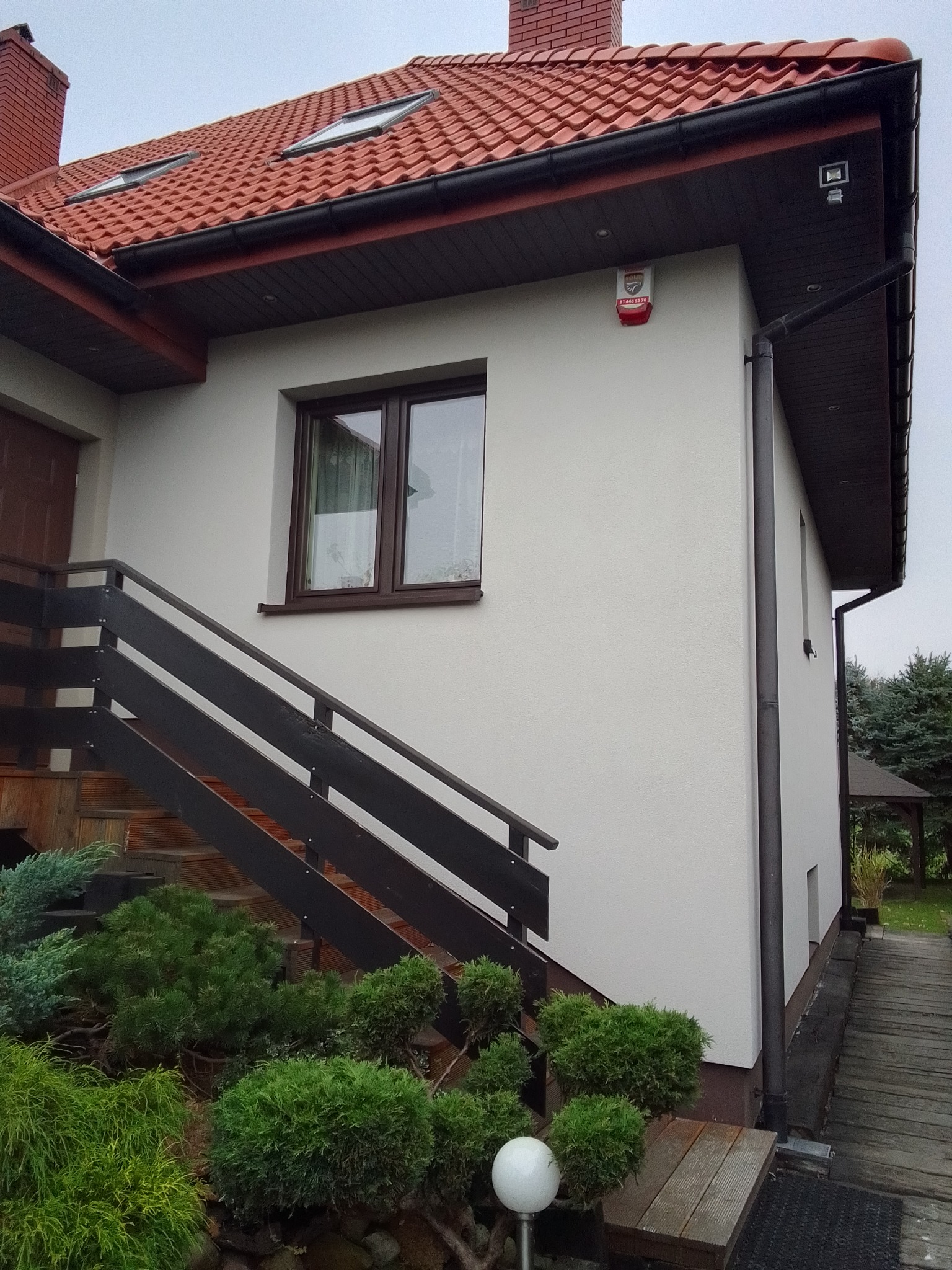 Fragment domu po remoncie elewacji: biała ściana z brązowymi oknami i schodami wejściowymi, czerwona dachówka, alarm na ścianie.