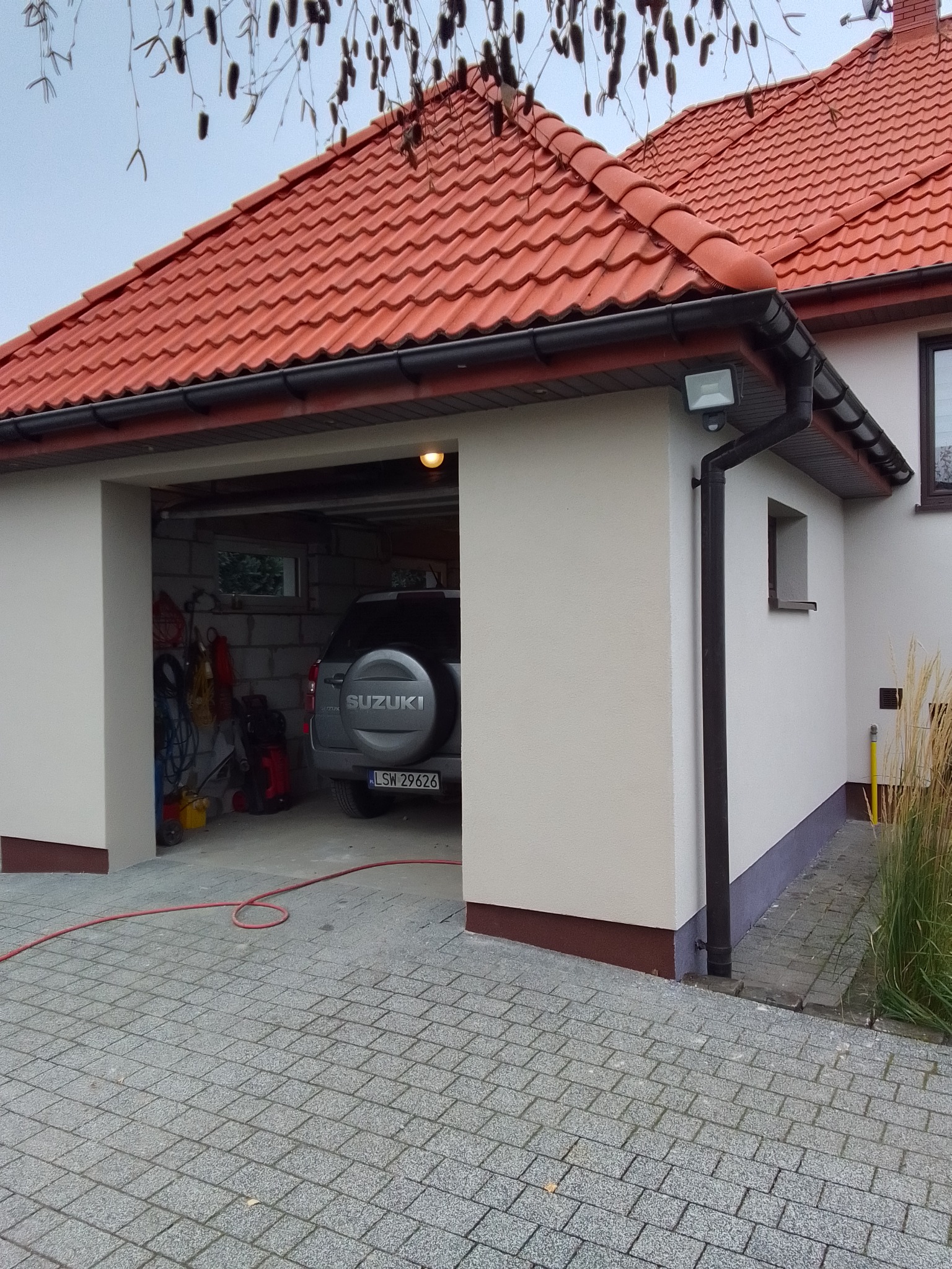 Nowoczesny garaż z czerwoną dachówką i jasną elewacją, widoczny samochód Suzuki w środku, kostka brukowa przed garażem.