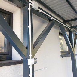 Volprim Instalacje Elektryczne Grzegorz Ciaranek - Białe rury elektryczne prowadzone po elewacji budynku, przymocowane do metalowej konstrukcji zadaszenia. Widoczna puszka elektryczna i czujnik ruchu.