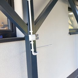 Volprim Instalacje Elektryczne Grzegorz Ciaranek - Zewnętrzna instalacja elektryczna w białych rurach PCV, zamontowana na metalowej konstrukcji budynku. Widoczna puszka elektryczna i fragment okna.