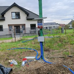 Volprim Instalacje Elektryczne Grzegorz Ciaranek - Zewnętrzna skrzynka elektryczna z gniazdami siłowymi na zielonym słupie, z niebieskimi rurami kablowymi, na tle domu jednorodzinnego i pochmurnego nieba.