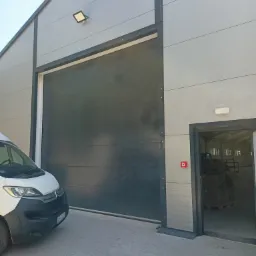 Szara brama przemysłowa segmentowa zamknięta, z widocznym fragmentem białego busa dostawczego i otwartymi drzwiami wejściowymi do hali, oświetlona lampą zewnętrzną. Na ścianie przy drzwiach...