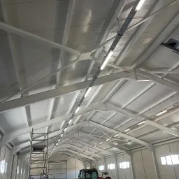 Wnętrze hali produkcyjnej z widoczną konstrukcją stalową dachu, oświetleniem liniowym LED zamontowanym na szynoprzewodach, oraz podnośnikiem koszowym Manitou i rozstawionym rusztowaniem.