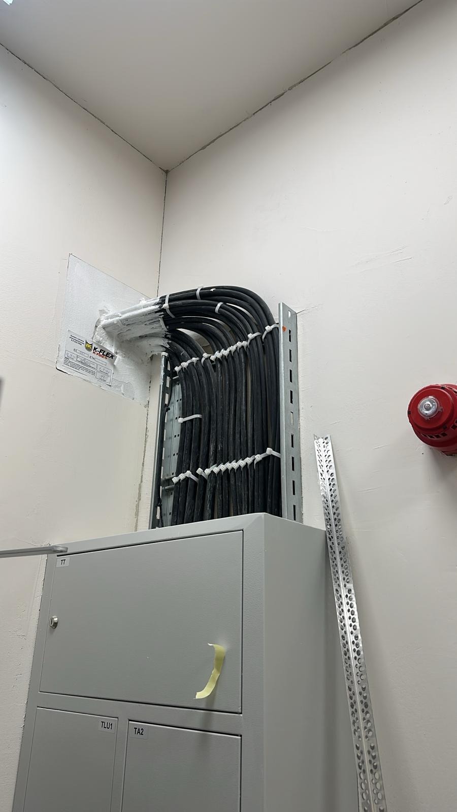Uporządkowane kable w metalowej korycie, zabezpieczone opaskami, wchodzące do szarej szafy elektrycznej. Widoczny czujnik dymu i fragment listwy montażowej.