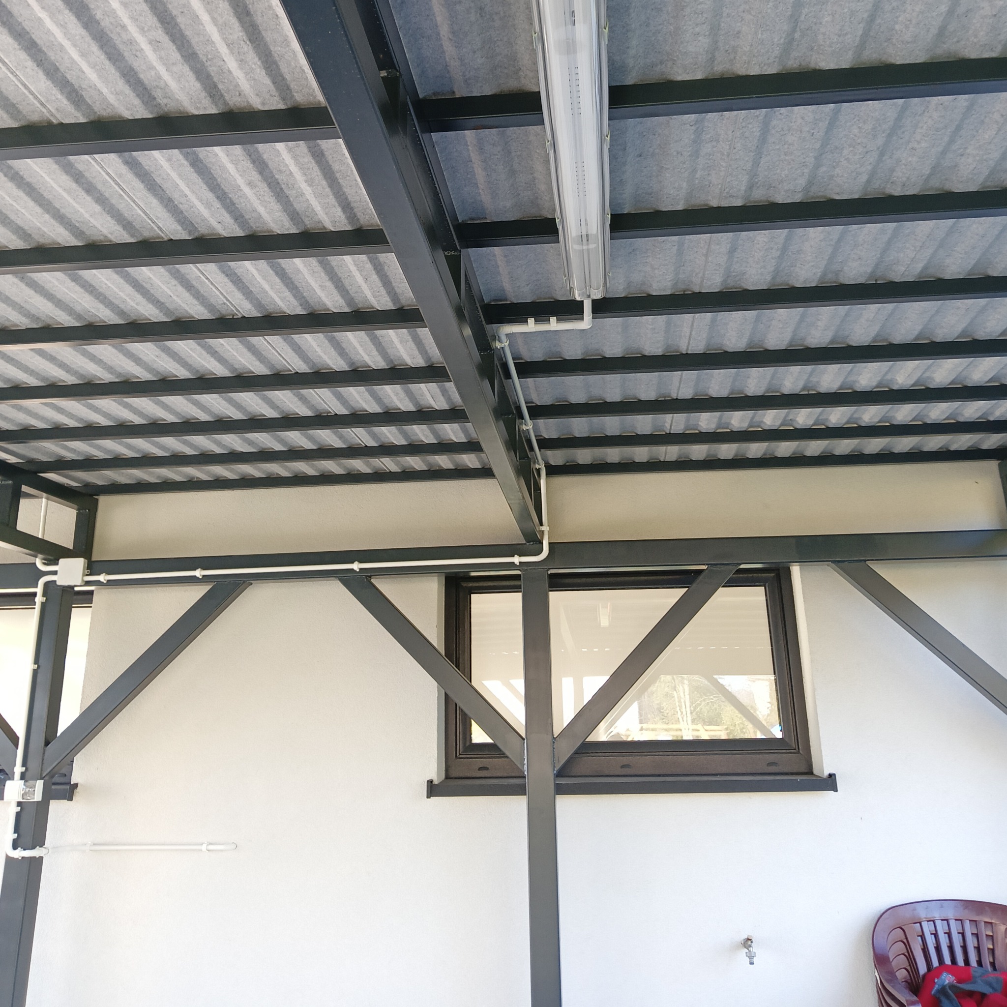 Zadaszenie tarasu z blachy trapezowej, oświetlone lampą LED. Widoczne rury instalacji elektrycznej i fragment okna. Konstrukcja stalowa, pomalowana na ciemny kolor.
