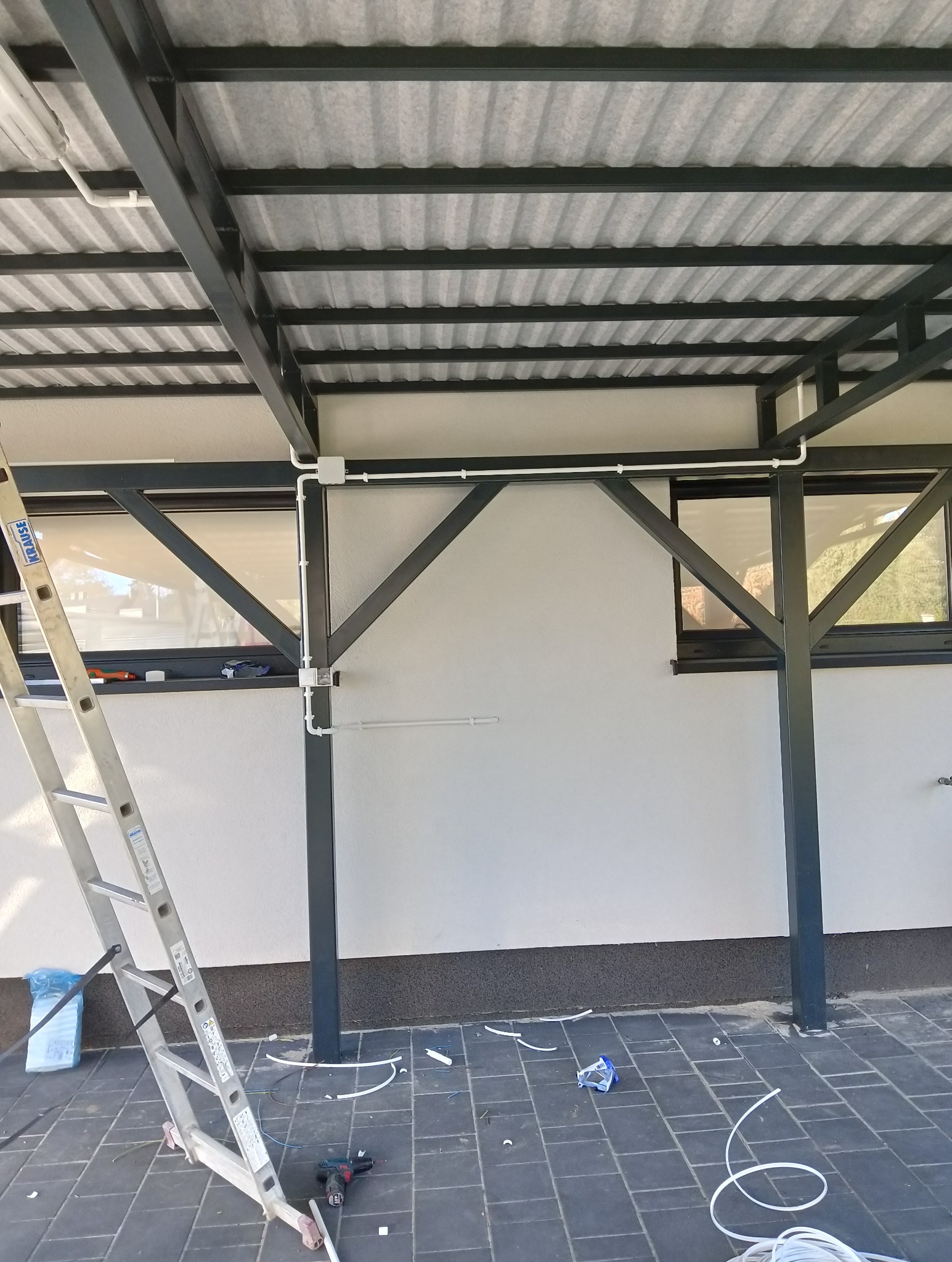 Białe rury instalacji elektrycznej zamontowane na ścianie pod zadaszeniem. Widoczna drabina, narzędzia i fragmenty przewodów na podłożu z kostki brukowej.