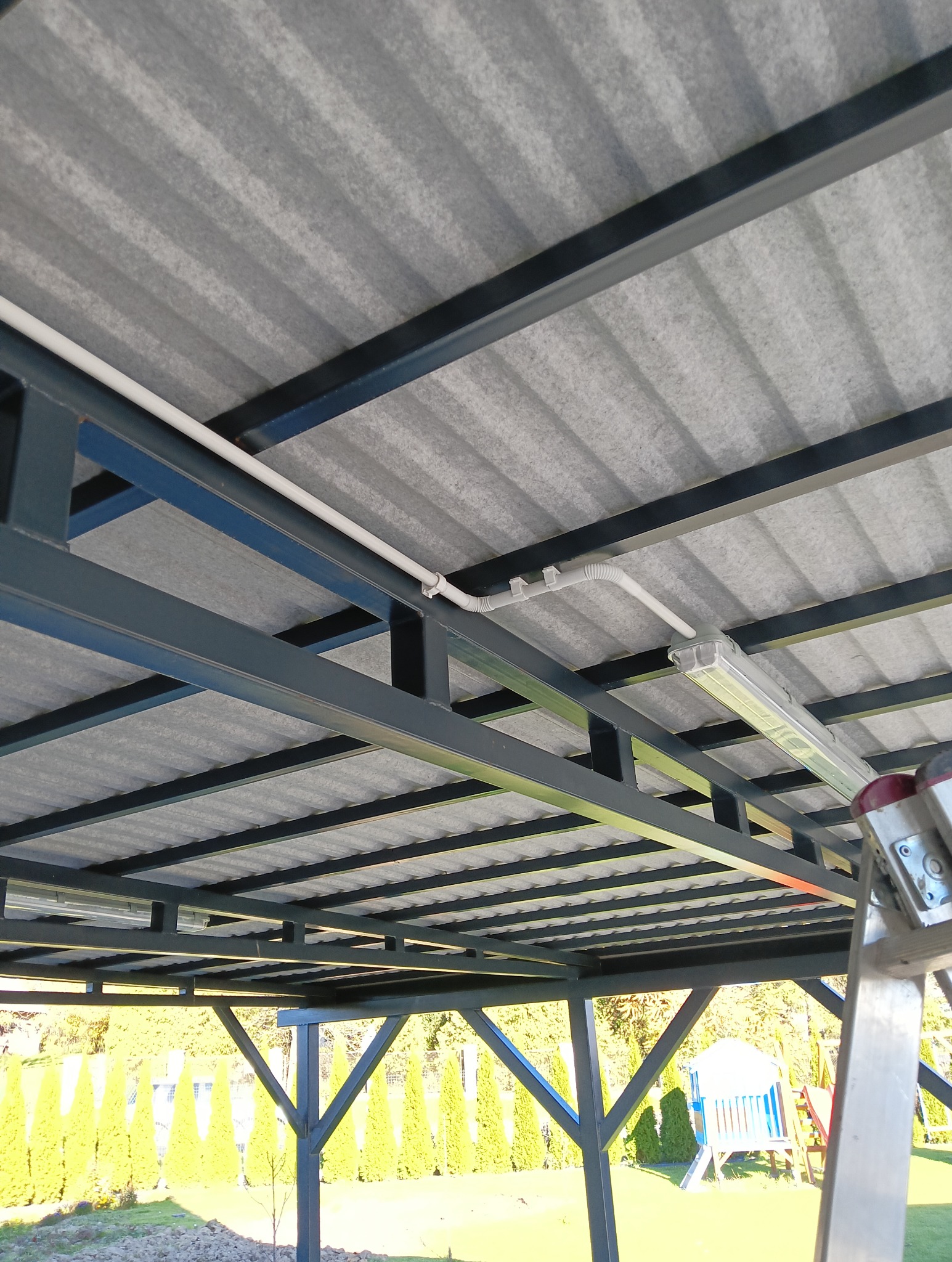 Szara konstrukcja z rurami i lampą na suficie, widok z dołu. Biała rura instalacji elektrycznej przymocowana do metalowej ramy. Oświetlenie pod zadaszeniem.