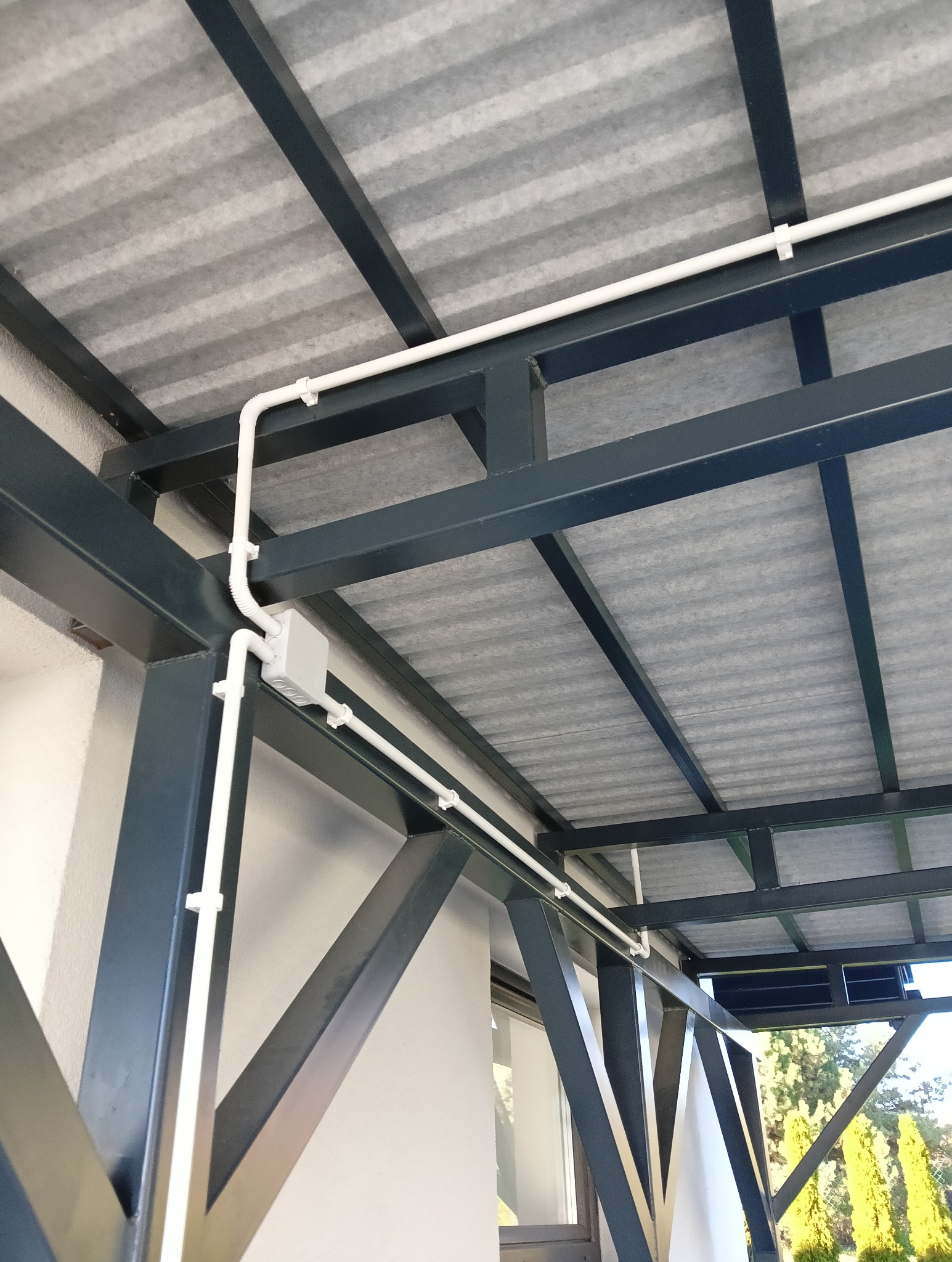 Białe rury elektryczne zamontowane pod szarym, falistym dachem na metalowej konstrukcji. Widoczna puszka instalacyjna i uchwyty mocujące rury. Ujęcie z dołu.