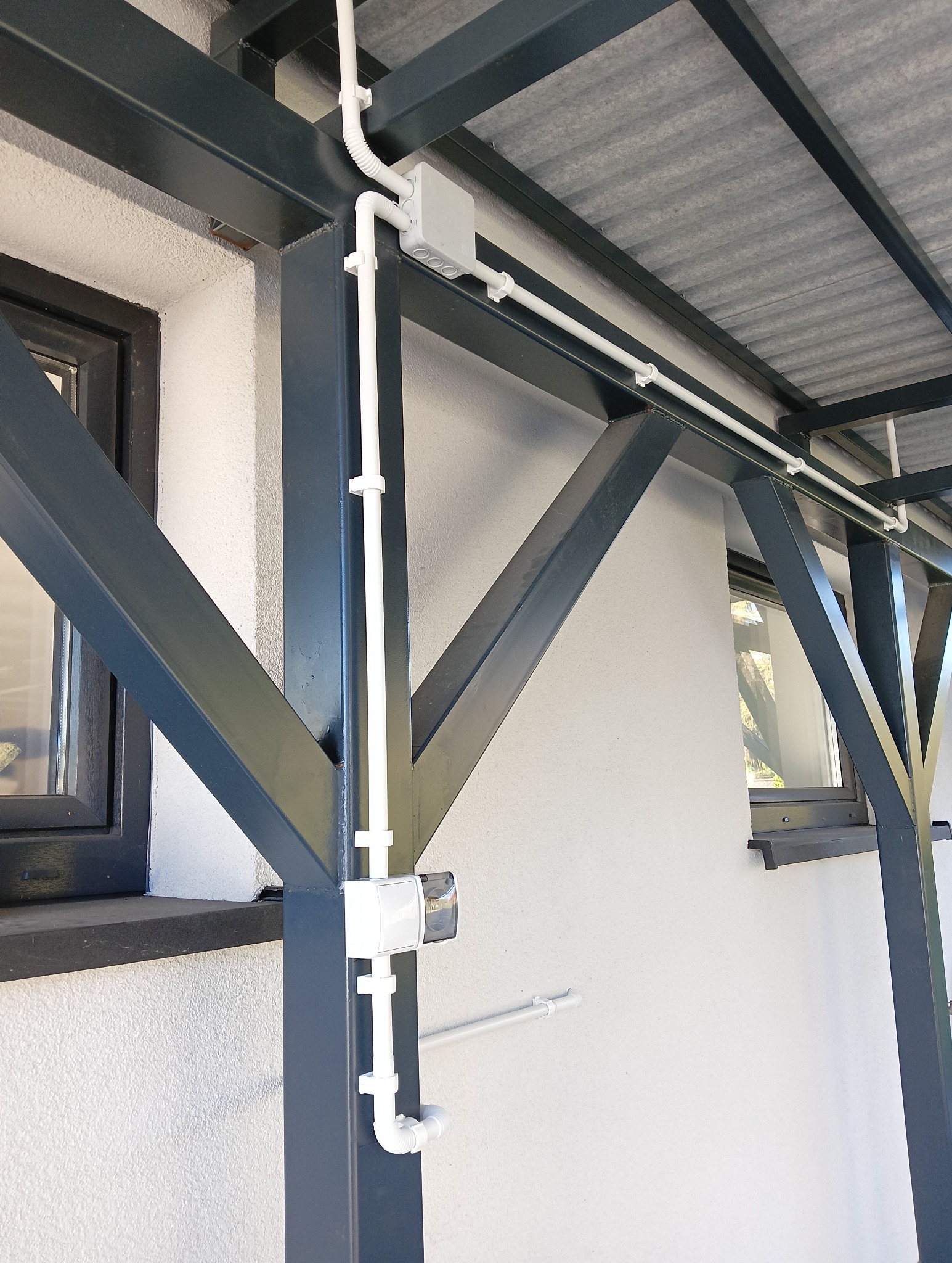 Białe rury elektryczne prowadzone po elewacji budynku, przymocowane do metalowej konstrukcji zadaszenia. Widoczna puszka elektryczna i czujnik ruchu.