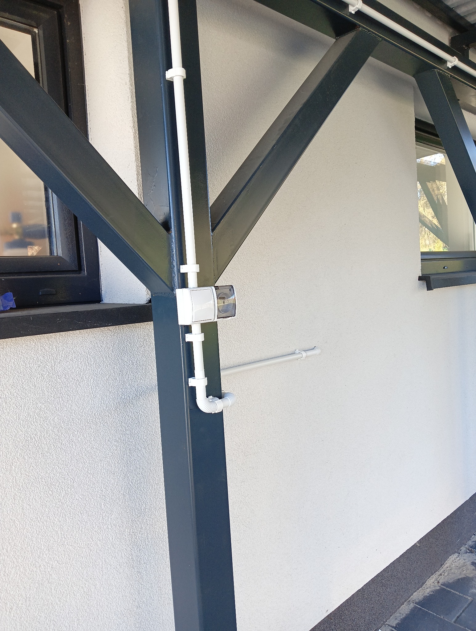 Zewnętrzna instalacja elektryczna w białych rurach PCV, zamontowana na metalowej konstrukcji budynku. Widoczna puszka elektryczna i fragment okna.
