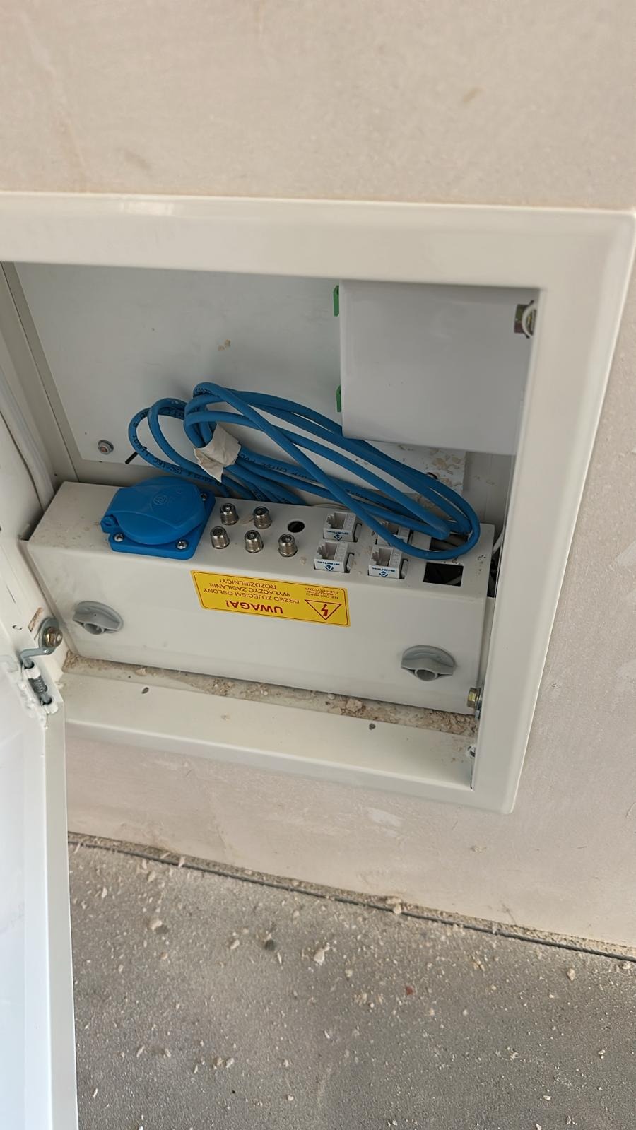 Otwarta skrzynka z gniazdami antenowymi, telefonicznymi i zasilającym, z niebieskim kablem sieciowym wewnątrz, zamontowana w ścianie. Widoczna naklejka ostrzegawcza.