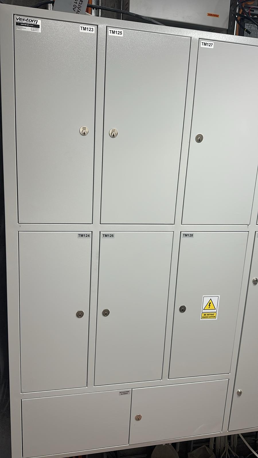 Szara rozdzielnica elektryczna z numerowanymi drzwiczkami i ostrzeżeniem 'Nie dotykać, porażenie elektryczne', widoczna instalacja w tle.