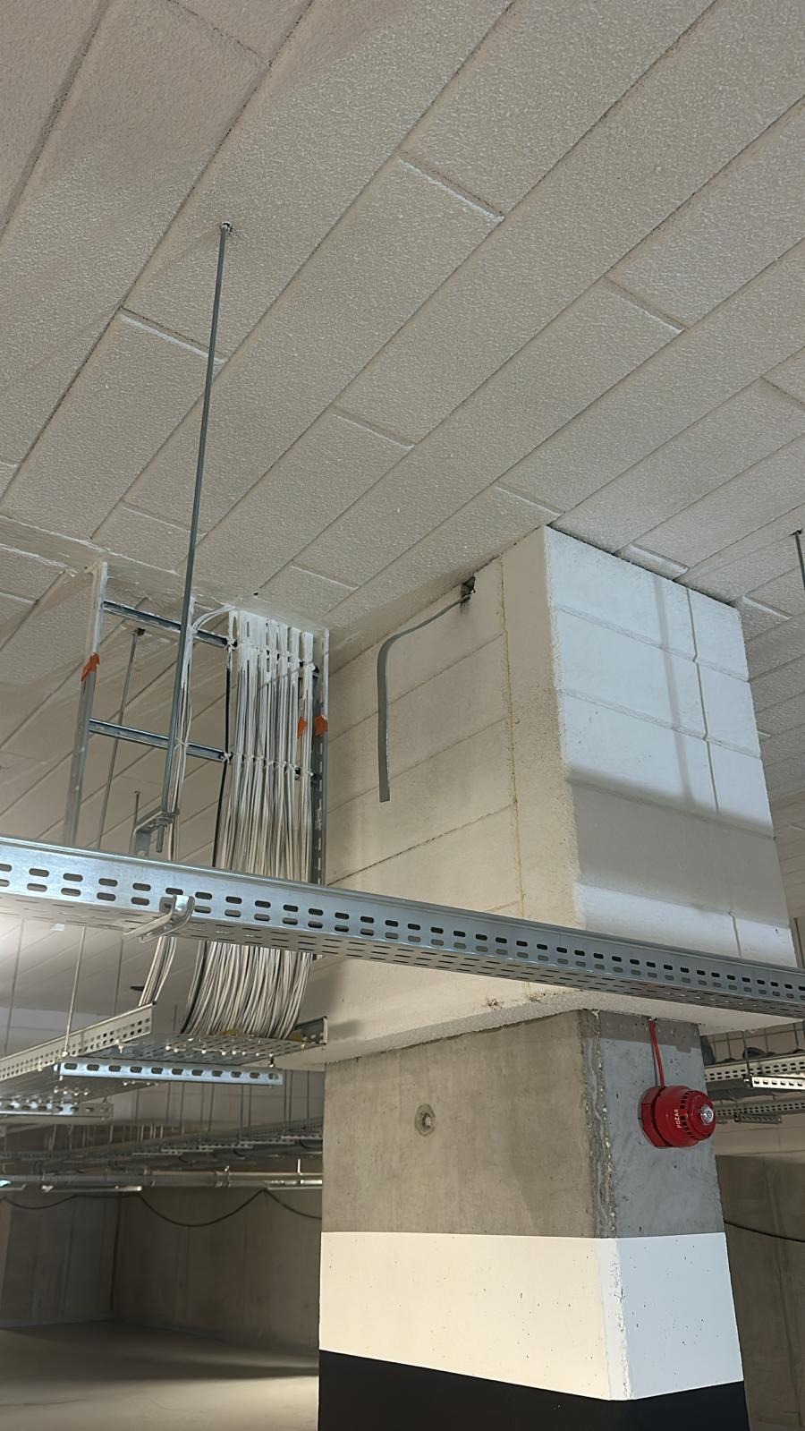 Kable w korytach zamocowane pod sufitem, widoczny czujnik przeciwpożarowy na filarze. Instalacja w nowym budynku.