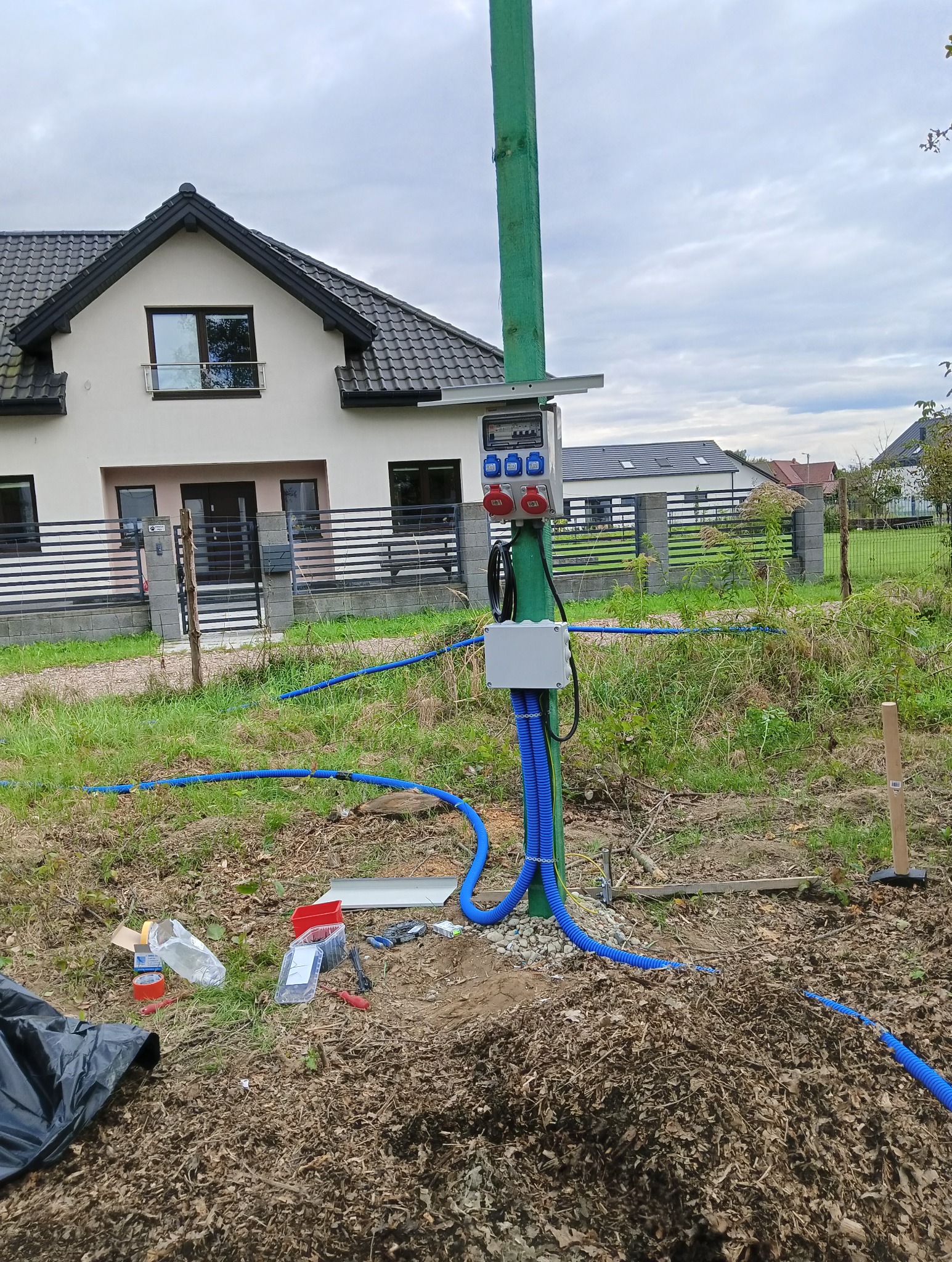 Zewnętrzna skrzynka elektryczna z gniazdami siłowymi na zielonym słupie, z niebieskimi rurami kablowymi, na tle domu jednorodzinnego i pochmurnego nieba.