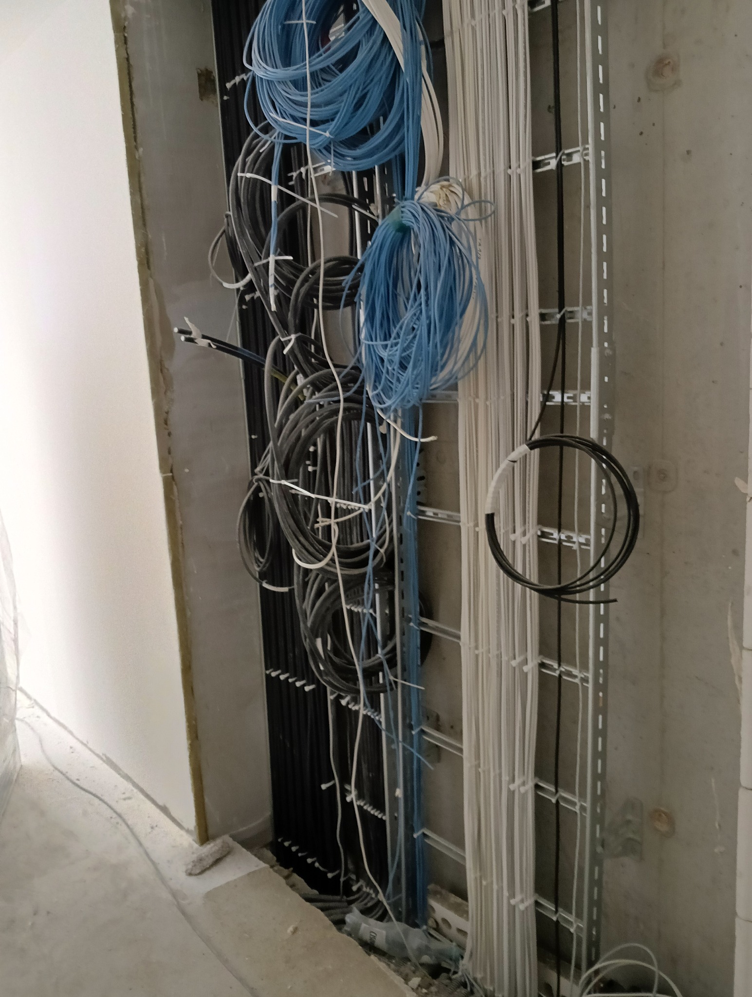 Wewnętrzna instalacja elektryczna w trakcie montażu: kable w różnych kolorach ułożone w metalowych korytkach na ścianie. Widoczne przewody czarne, niebieskie i białe.
