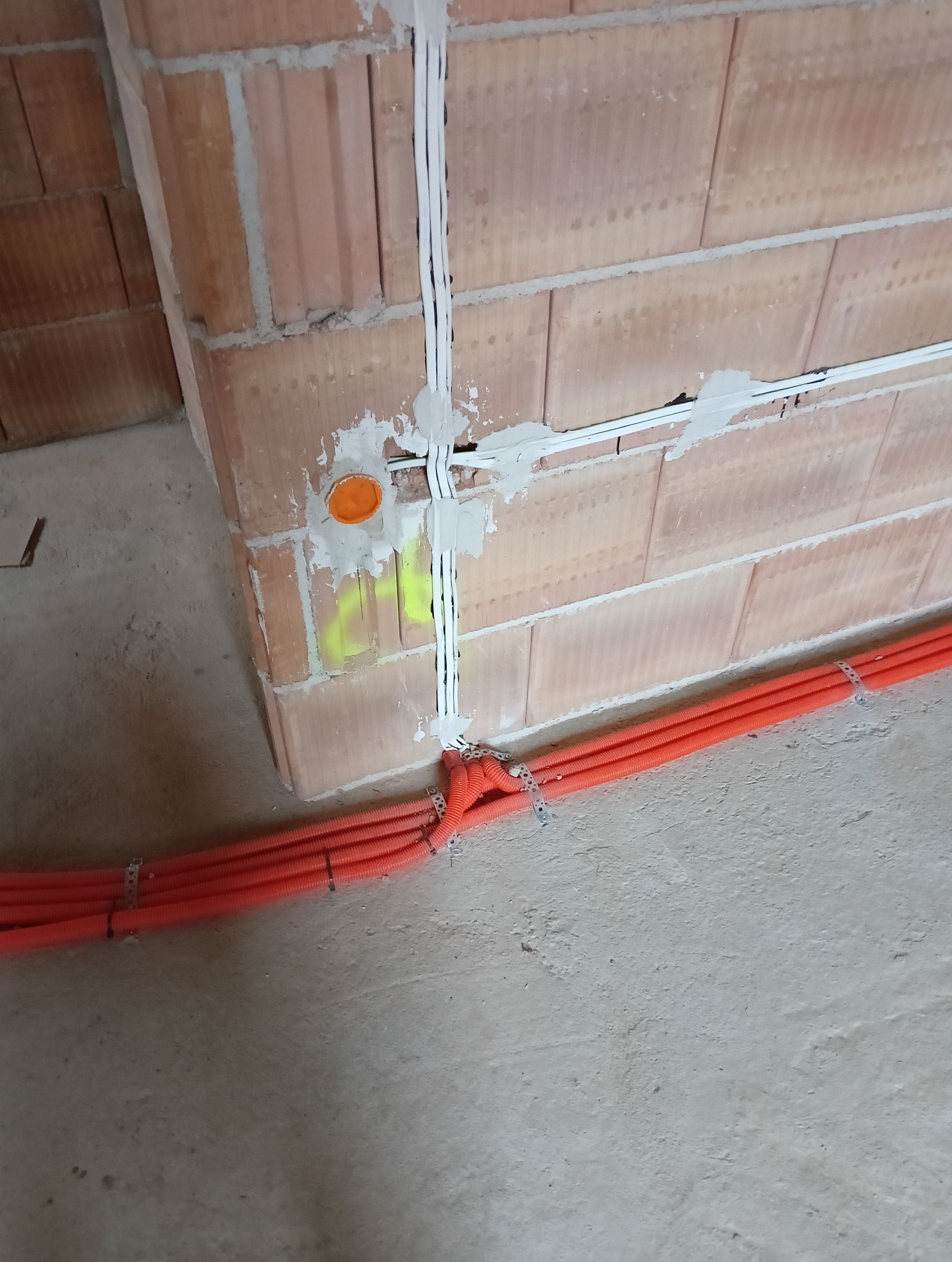Białe przewody i pomarańczowe rury w ścianie z cegły dziurawki, zabezpieczone uchwytami. Instalacja elektryczna w trakcie realizacji w Krakowie.