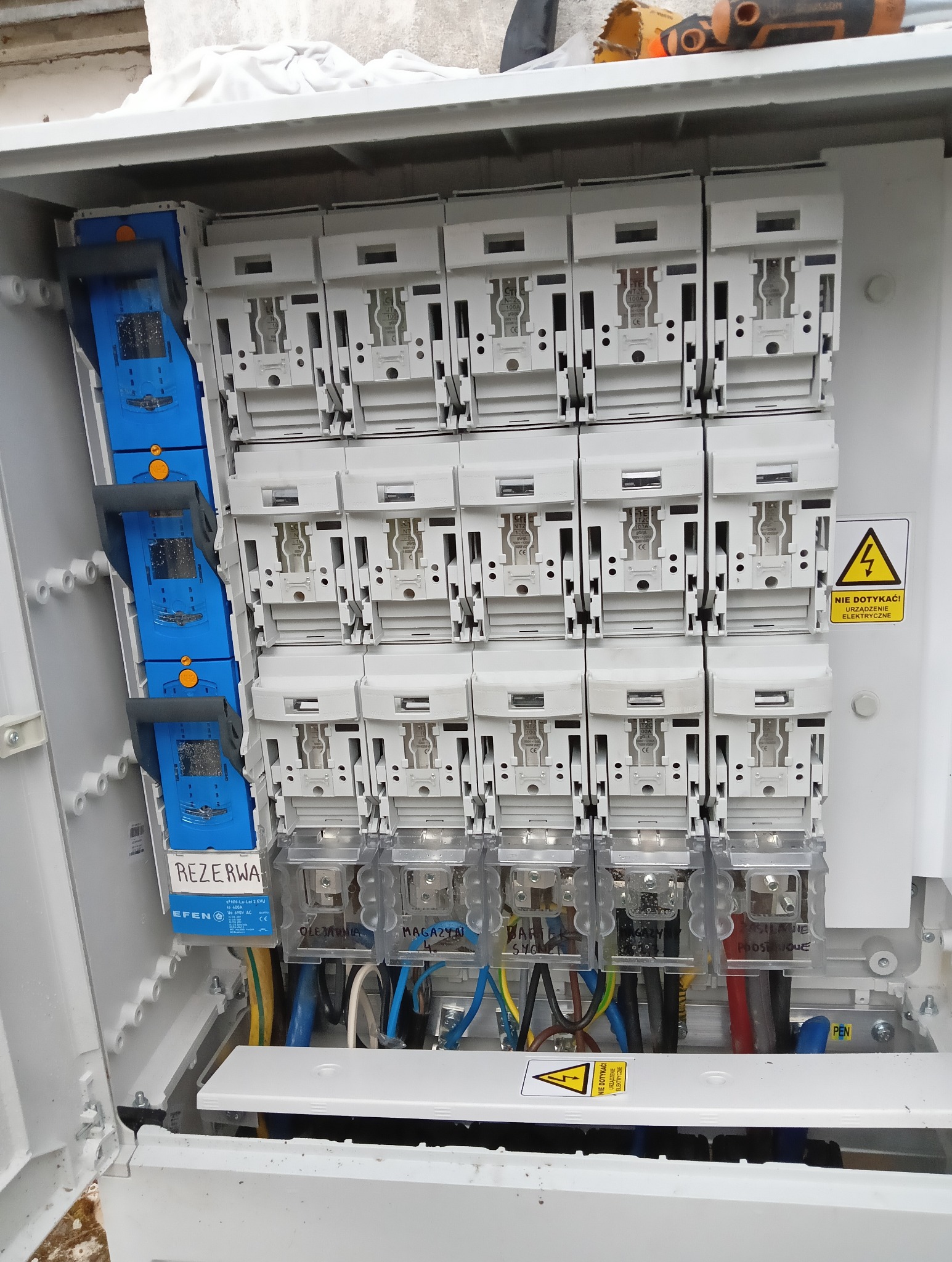 Otwarta skrzynka elektryczna z bezpiecznikami i okablowaniem. Widoczne oznaczenia na bezpiecznikach i ostrzeżenie o napięciu. Ujęcie z bliska, ukazujące detale instalacji.