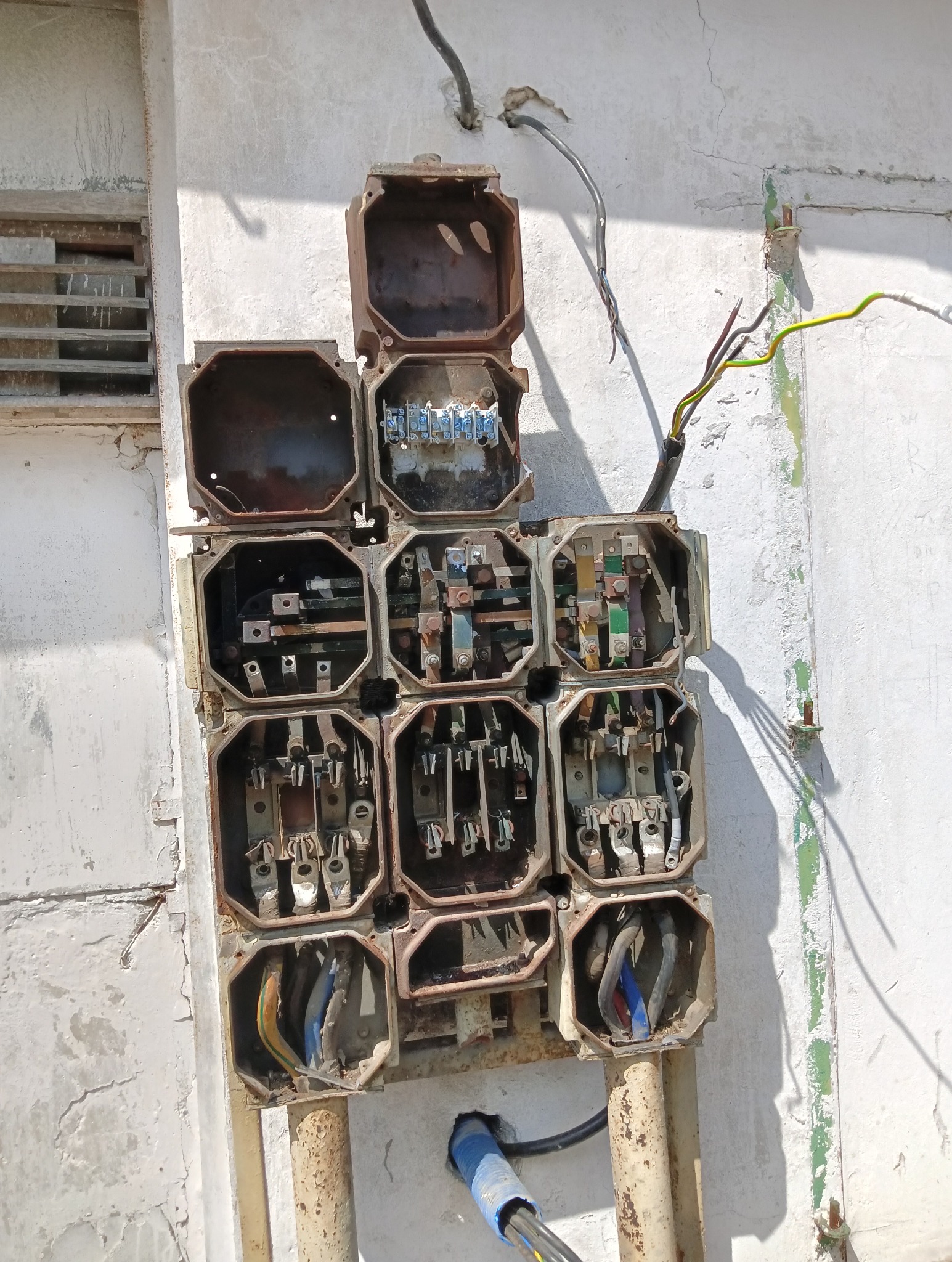 Otwarta, skorodowana skrzynka elektryczna z widocznymi przewodami i elementami łączeniowymi, zamontowana na pomalowanej ścianie budynku. Widoczne ślady korozji i zużycia.