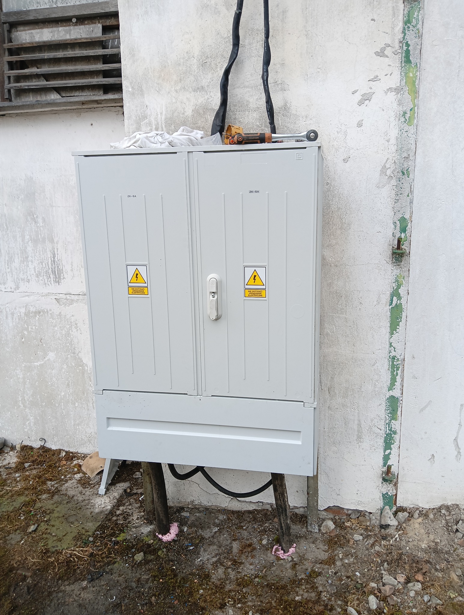 Szara skrzynka elektryczna ZK-5A z ostrzeżeniem o napięciu, zamontowana na zewnątrz budynku. Na górze leży klucz płaski. Widoczne kable i fragment ściany.