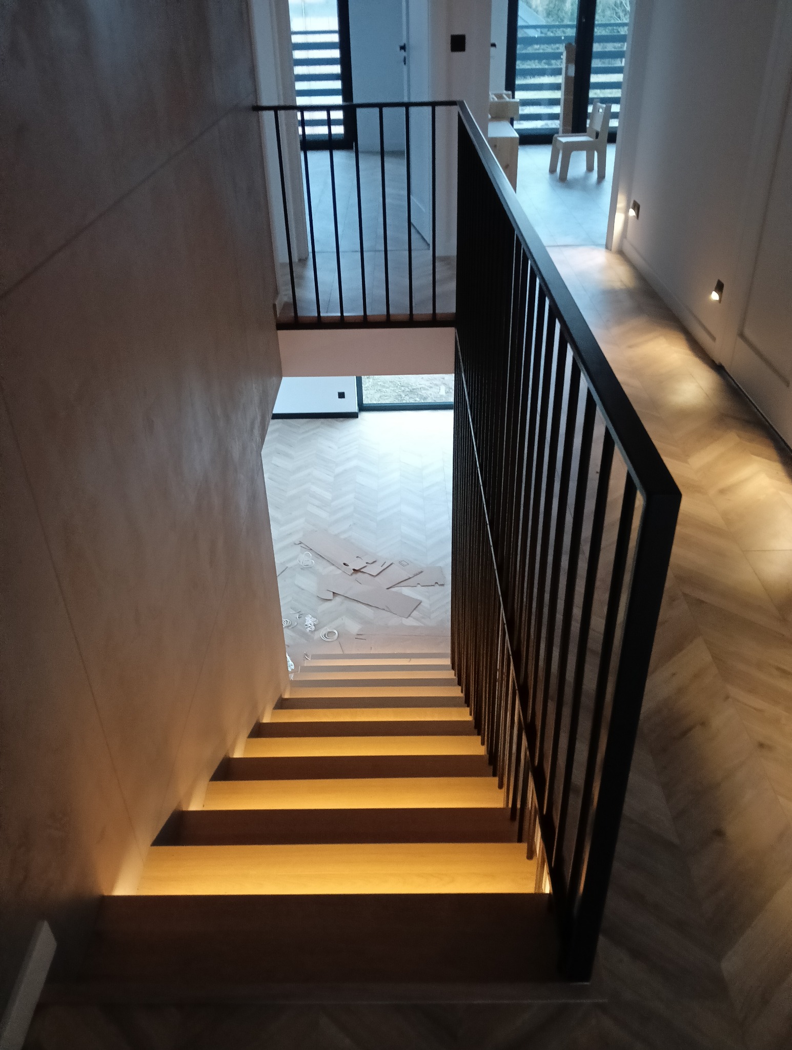 Schody z podświetleniem LED i czarną balustradą w nowoczesnym domu. Widok z góry na parkiet w jodełkę i elementy wykończeniowe na podłodze.