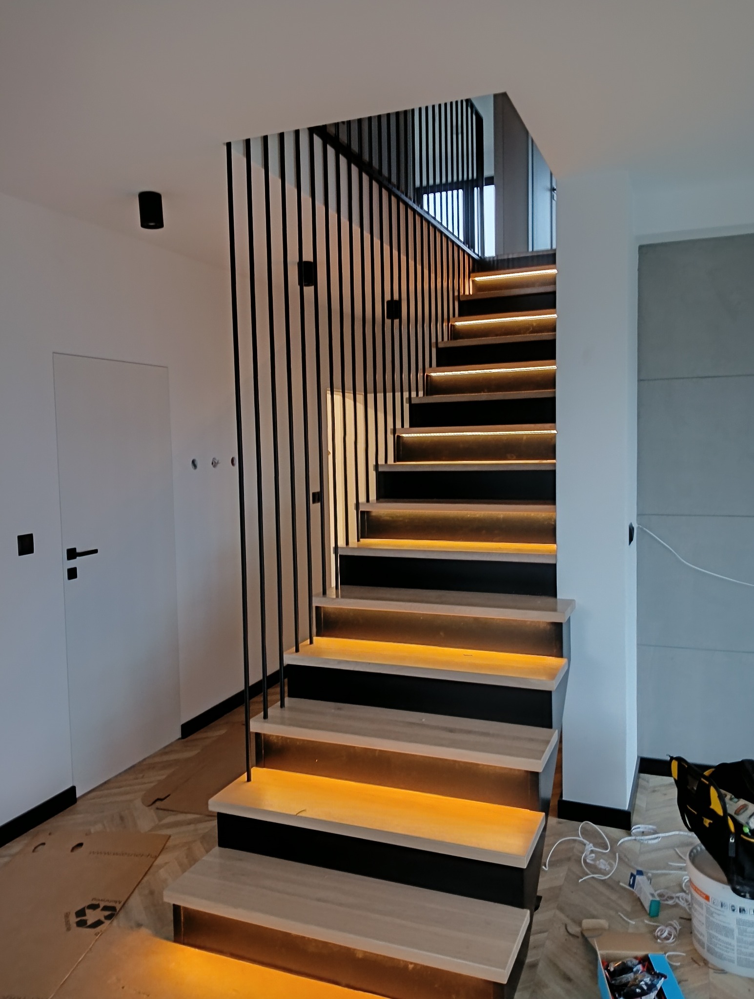 Nowoczesne schody z podświetleniem LED, czarne barierki, jasne drewno. Widoczne narzędzia i materiały, sugerujące etap montażu oświetlenia.