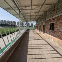 Zadaszona konstrukcja stalowa z poręczami, ceglana ściana budynku gospodarczego i widok na zielone pole w tle.