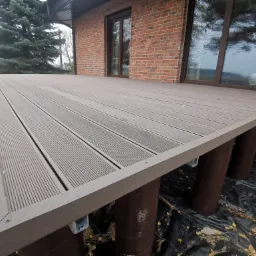 Nowy taras z kompozytowych desek w odcieniach brązu, wsparty na okrągłych słupach, z widoczną ceglaną ścianą budynku w tle.