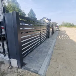Nowoczesna, pozioma, przesuwna brama wjazdowa w kolorze antracytowym, z widocznymi poziomymi panelami i solidnymi słupkami, zamontowana na posesji z kostką brukową i ogrodzeniem z siatki.