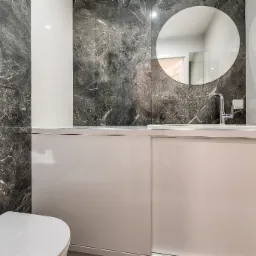 Nowoczesna łazienka z umywalką w zabudowie, okrągłym lustrem i marmurowymi płytkami ściennymi w odcieniach szarości i bieli, widoczny fragment toalety.