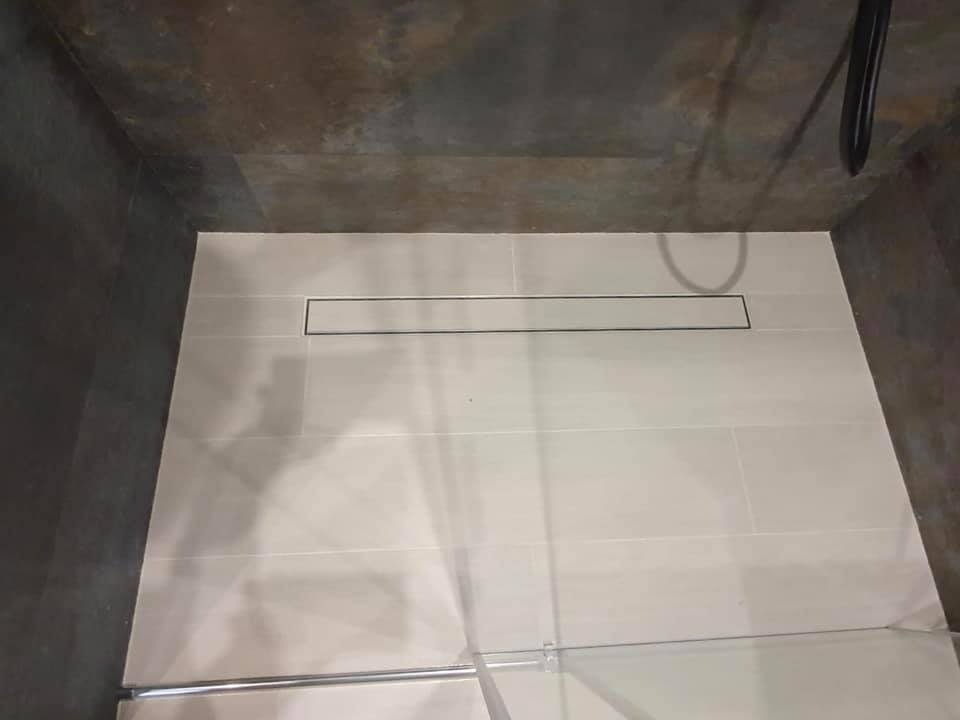 Nowoczesny odpływ liniowy w jasnej, wykafelkowanej kabinie prysznicowej z ciemnymi ścianami i sufitem o teksturze kamienia.