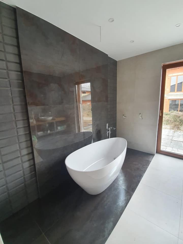 Nowoczesna łazienka z wolnostojącą białą wanną o owalnym kształcie, ciemnoszara ściana z płytek o fakturze 3D po lewej, duża płyta imitująca kamień na ścianie, okno z drewnianą ramą po prawej...