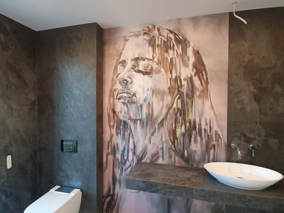 Nowoczesna łazienka z umywalką nablatową, toaletą i dużą grafiką kobiety na ścianie, wykonana w ciemnej kolorystyce z efektem kamienia.