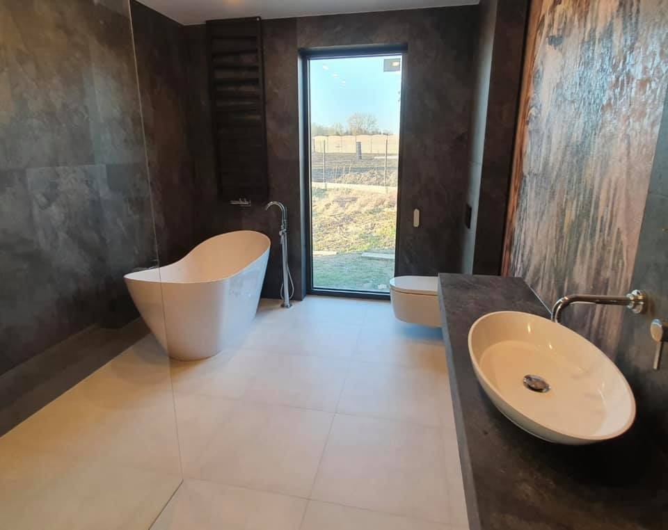 Nowoczesna łazienka z wolnostojącą wanną, umywalką nablatową na ciemnym blacie i dużym oknem z widokiem na zewnątrz, ściany wykończone w ciemnym, strukturalnym tynku.