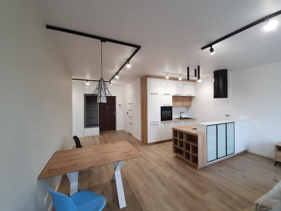 Jasne wnętrze mieszkania po remoncie: drewniana podłoga, białe ściany, nowoczesna zabudowa kuchenna z wyspą, czarny okap cylindryczny, oświetlenie szynowe z geometryczną lampą wiszącą...