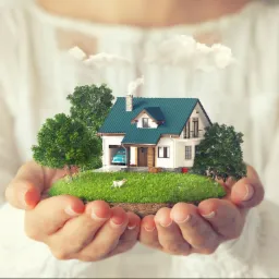 Model domu z zielonym dachem, trawnikiem, drzewami, rowerem i psem, trzymany w dłoniach na tle białej koszuli.