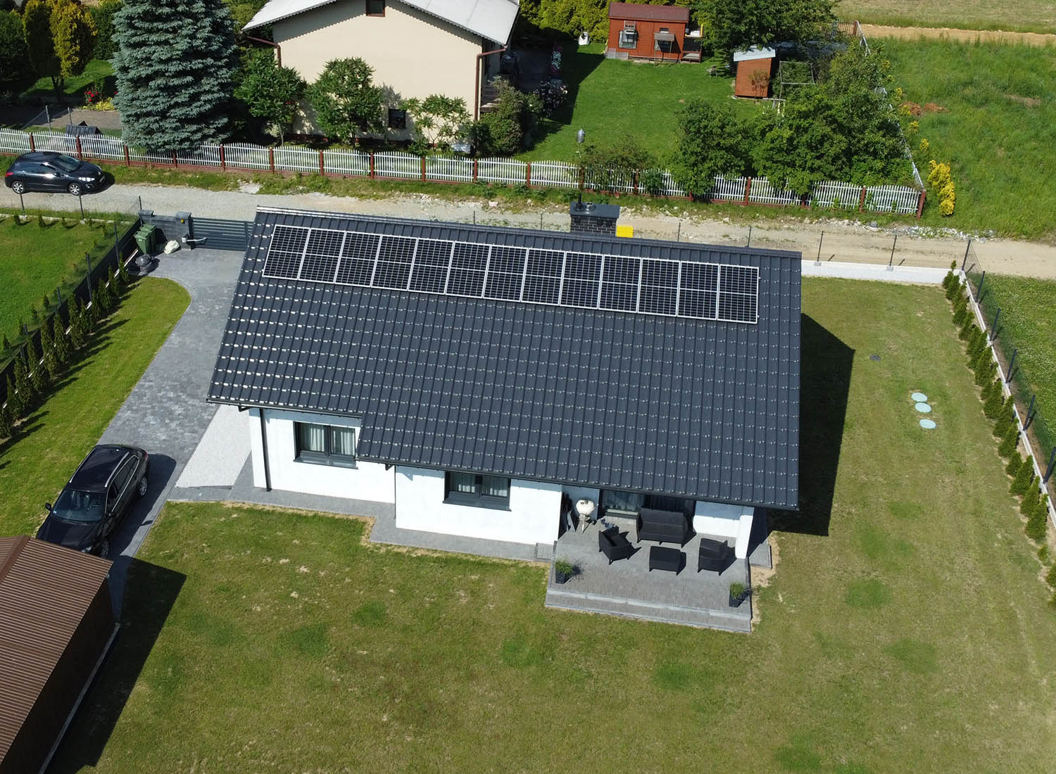 Widok z lotu ptaka na dom jednorodzinny z panelami słonecznymi na dachu, szary dach z blachy trapezowej, biała elewacja, zestaw mebli ogrodowych na tarasie, zadbany trawnik i podjazd z kostki...