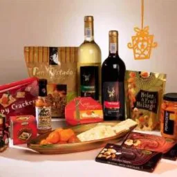 Bogato zaopatrzony zestaw prezentowy z winem białym i czerwonym, krakersami, czekoladą, konfiturami i dekoracyjną zawieszką w kształcie kokardy, umieszczony na beżowym tle.