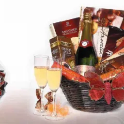 Elegancki kosz upominkowy z butelką wina musującego, słodyczami i dekoracjami, obok mniejszy kosz zapakowany w celofan, dwa kieliszki wypełnione napojem.