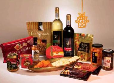 Bogato zaopatrzony zestaw prezentowy z winem białym i czerwonym, krakersami, czekoladą, konfiturami i dekoracyjną zawieszką w kształcie kokardy, umieszczony na beżowym tle.