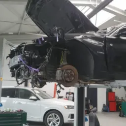Czarny samochód hybrydowy na podnośniku w warsztacie, z uszkodzonym przodem i otwartą maską, widoczne elementy silnika i zawieszenia, obok biały SUV z uszkodzonym tyłem, skrzynka z narzędziami...