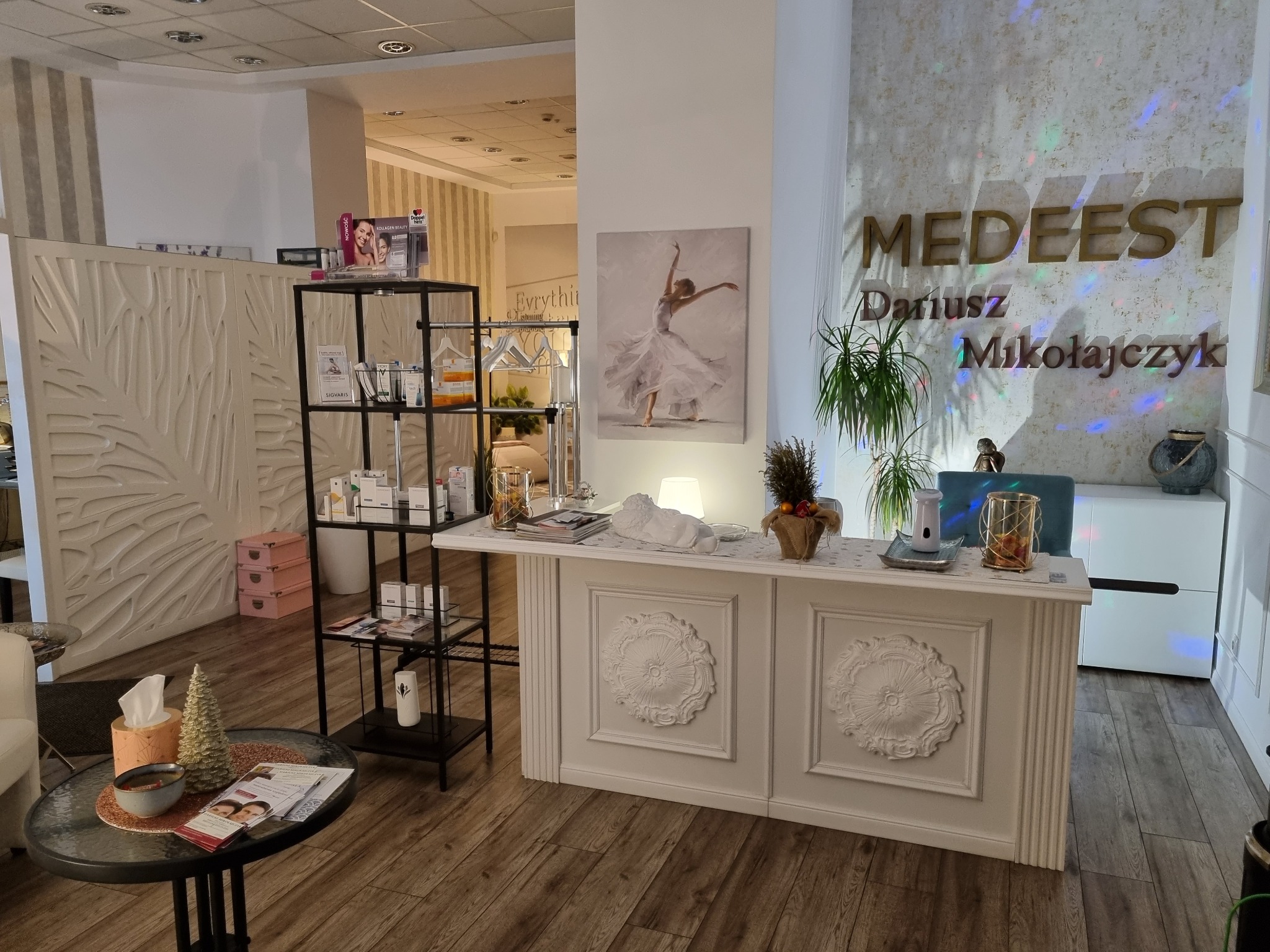 Jasne, przestronne wnętrze recepcji z białym biurkiem ozdobionym rozetami, półkami z produktami i dekoracjami, w tle obraz baletnicy i logo firmy.